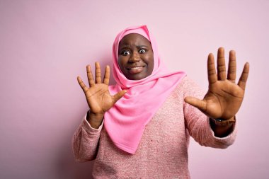 Genç Afro-Amerikan artı cüsseli kadın soyutlanmış pembe arka plan üzerine Müslüman başörtüsü takıyor korku ifadesinden korkuyor ve dehşete düşüyor hareketlerini elleriyle durduruyor, şok içinde bağırıyor. Panik konsepti.