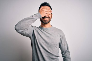 Sakallı, sıradan bir süveter giyen, beyaz arka planda dikilen, gülümseyen ve yüzünde şaşkınlıktan gözleri kapayan genç yakışıklı bir adam. Kör kavram.