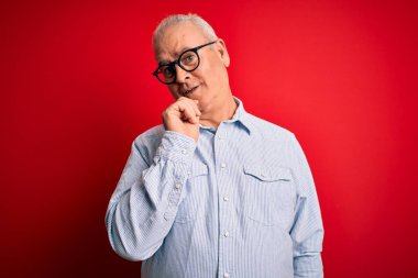 Orta yaşlı, beyaz, sıradan çizgili tişört ve kırmızı arka planda gözlük takan yakışıklı adam kameraya kollarını kavuşturup gülümseyen ve çenesini kaldıran adama bakıyor. Olumlu düşünüyorum..