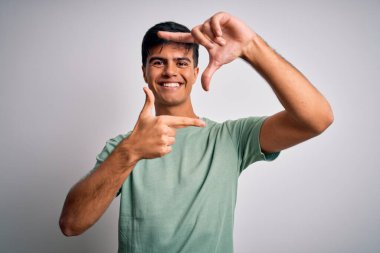 İzole edilmiş beyaz arka plan üzerinde sıradan tişörtler giyen genç yakışıklı adam gülümsüyor ve mutlu yüzlü elleri ve parmakları olan bir çerçeve yapıyor. Yaratıcılık ve fotoğrafçılık kavramı.