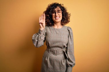 Sarı arka plan üzerinde duran elbise ve gözlük takan güzel Arap iş kadını dört numaralı parmaklarıyla işaret ederken kendinden emin ve mutlu bir şekilde gülümsüyor..