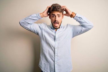 Beyaz arka plan üzerinde duran çizgili tişörtlü, sakallı, genç, yakışıklı adam elleri kafasında çılgın, korkmuş ve ağzı açık şoktan şaşırmış.