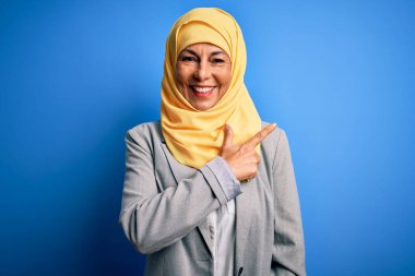 Orta yaşlı esmer iş kadını, geleneksel Müslüman tesettüre bürünmüş, mavi arka planda neşeli, yüzünde bir gülümseme, mutlu ve doğal bir ifadeyle elini ve parmağını kaldırmış.