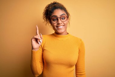 Sarı arka planda süveter ve gözlük takan güzel Afro-Amerikan kızı bir numaralı parmağını gösterirken kendine güvenen ve mutlu bir şekilde gülümsüyor..