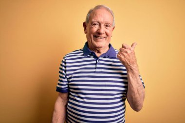 Gri saçlı, donanma çizgili tişört giyen, sarı arka planda dikilen, gülümseyen ve baş parmağıyla yan tarafı gösteren son sınıf öğrencisi..