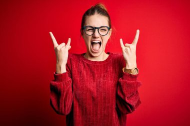Genç, güzel, kızıl saçlı bir kadın izole edilmiş kırmızı arka plan üzerine günlük süveter giyip çılgın bir ifadeyle el kaldırarak rock sembolü yapıyor. Müzik yıldızı. Ağır müzik konsepti.