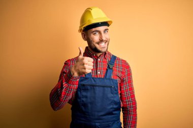 İnşaatçı üniformalı genç bir adam ve sarı izole edilmiş arka plan üzerine güvenlik kaskı takarak mutlu bir el hareketi yapıyor. Kameraya bakıp başarıyı gösteren ifade onaylanıyor.