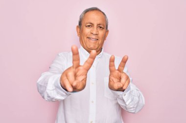 Üst düzey yakışıklı, gri saçlı, zarif tişörtlü, izole edilmiş pembe arka planda gülümseyen, parmaklarını zafer işareti yaparken gösteren kameraya bakan adam. İki numara..