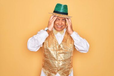 Orta yaşlı, kır saçlı, Brezilya karnavalı kıyafeti giyen yaşlı bir adam sarı arka planda çaresiz ve stresli baş ağrısından muzdarip, çünkü ağrı ve migren. Ellerini başının üstüne koy..