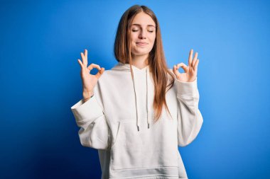 Genç, güzel, kızıl saçlı sporcu kadın izole edilmiş mavi arka plan üzerine kazak giyiyor rahatla ve gözleri kapalı, parmaklarıyla meditasyon hareketi yaparken gülümse. Yoga konsepti.