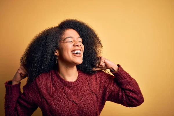 Young beautiful african american woman with afro hair standing over yellow isolated background very happy and excited doing winner gesture with arms raised, smiling and screaming for success. Celebration concept.