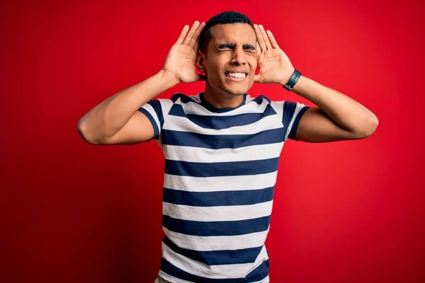 Handsome african american man wearing casual striped t-shirt standing over red background Trying to hear both hands on ear gesture, curious for gossip. Hearing problem, deaf