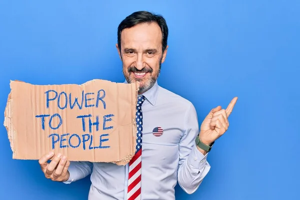 Middle age business man wearing usa tie holding banner with power to the people message smiling happy pointing with hand and finger to the side