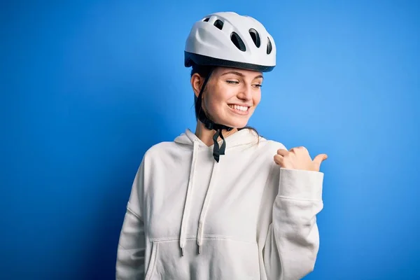 若いです美しいです赤毛サイクリスト女性身に着けていますバイクヘルメット上の隔離された青の背景笑顔で幸せな顔見ますと指で側面を指していますアップ — ストック写真