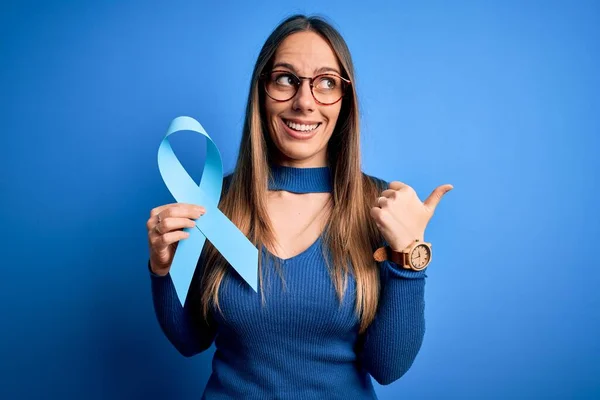 Young blonde woman with blue eyes holding colon cancer awareness blue ribbon pointing and showing with thumb up to the side with happy face smiling