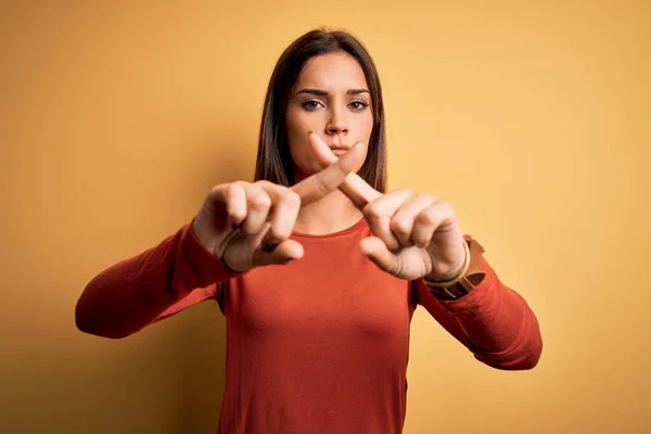 年轻美丽的黑发女人穿着休闲装站在黄色背景上 用手指交叉表示拒绝 做阴性手势 — 图库照片