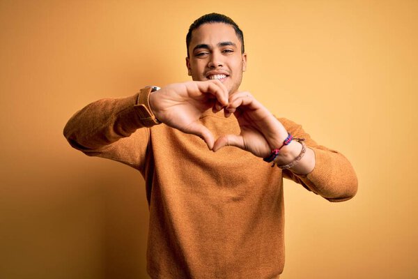 Young brazilian man wearing casual sweater standing over isolated yellow background smiling in love doing heart symbol shape with hands. Romantic concept.