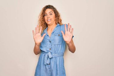 Mavi gözlü genç bir kadın beyaz arka planda sıradan bir kot elbise giyiyor. Korkudan dehşete düşmüş ve korkmuş. El hareketlerini bırak, şok içinde bağır. Panik konsepti.