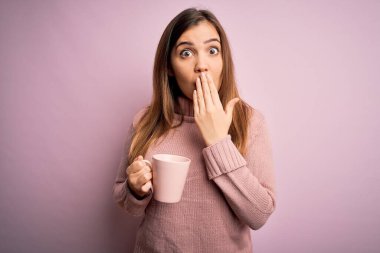 Sarışın genç bir kadın pembe izole edilmiş arka planda bir fincan kahve içiyor. Ağzı kapalı, utancından ağzı açık kalmış, korkunun dışa vurumu, sessizlikten korkması, gizli konsept.