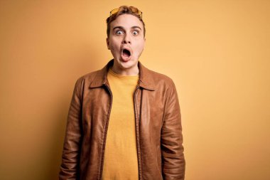 Genç, yakışıklı, deri ceket giyen, izole edilmiş sarı bir arka plan giyen, şaşkınlık ve şaşkınlık içinde, korku ve heyecan dolu bir yüz ifadesi olan, genç, kızıl saçlı adam..