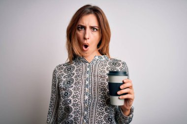 Genç, güzel esmer kadın beyaz arka planda bir bardak kahve içiyor. Şaşırmış bir yüzle korkmuş ve korku ifadesinden heyecanlanmış.