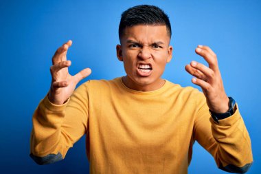 Genç, yakışıklı, latin bir adam, sarı günlük süveter giyiyor, mavi arka planda, öfkeden çıldırmış bağırıyor, elleri boğazını sıkmaya çalışıyor, deli gibi bağırıyor.