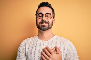 Sakallı, sıradan tişörtlü, sarı arka planda gözlüklü, göğsünde elleri kapalı, yüzünde minnet ifadesi olan yakışıklı bir adam. Sağlık kavramı.