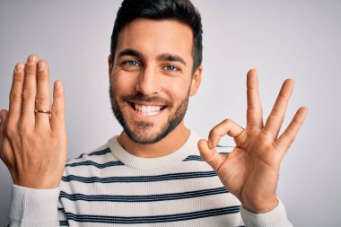 Sakallı yakışıklı bir adam beyaz arka planda evlilik yüzüğü taktığını gösteriyor. Parmaklarıyla imza atıyor, mükemmel bir sembol.