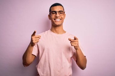 Yakışıklı Afro-Amerikan erkeği pembe arka plan üzerine günlük tişört ve gözlük takıp mutlu ve komik yüzlü parmakları kameraya doğrultuyor. İyi enerji ve titreşimler.