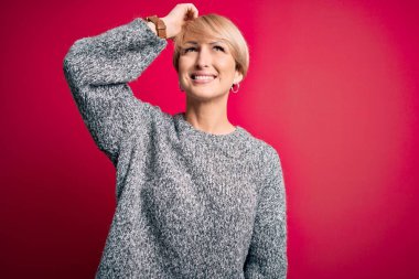 Modern kısa saçlı, pembe arka planda rahat süveter giyen, kendine güvenen, el hareketleriyle saçlarına dokunan çekici ve şık görünümlü genç sarışın kadın.