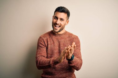 Genç, yakışıklı bir adam, günlük süveter giyer, izole beyaz arka planda durur, alkışlar, alkışlar ve mutlu, mutlu, gururlu eller bir arada.