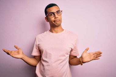 Yakışıklı Afro-Amerikalı adam pembe arka planda günlük tişört ve gözlük takıyor. Hiçbir şeyden habersiz ve kolları açık, hiçbir fikri yok..