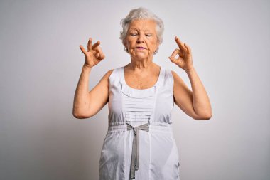 Son sınıf güzeli, gri saçlı, beyaz arka plan üzerine yazlık elbise giyen, gözleri kapalı, meditasyon yaparken parmaklarıyla gülümseyen bir kadın. Yoga konsepti.