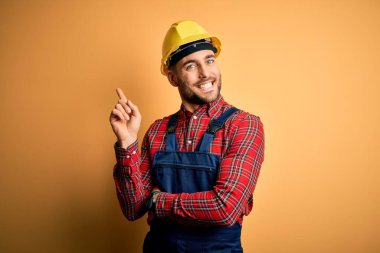 İnşaat üniforması giyen ve sarı izole edilmiş arka planda güvenlik kaskı takan genç bir müteahhit yüzünde büyük bir gülümsemeyle kameraya el ve parmak ile bakıyor..