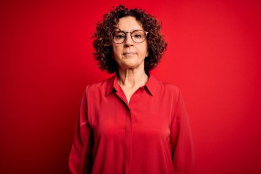 Orta yaşlı, kıvırcık saçlı, rahat gömlekli ve kırmızı arka planda gözlüklü güzel bir kadın. Yüzünde ciddi bir ifadeyle rahatlamış. Basit ve doğal bir şekilde kameraya bakıyorsun..