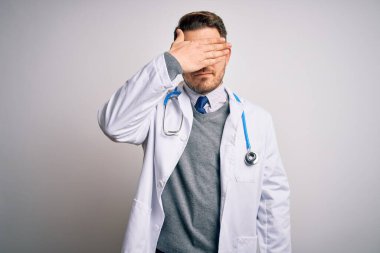 Mavi gözlü genç doktor tıbbi ceket giyiyor ve steteskop ile izole edilmiş arka planda, elleri ile gözlerini kapatıyor, ciddi ve üzgün görünüyor. Görme, saklama ve reddetme konsepti