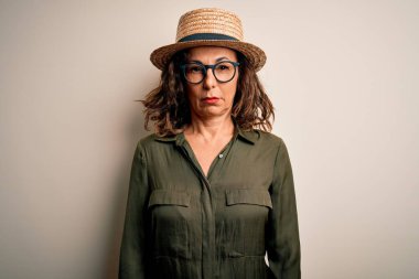 Orta yaşlı esmer kadın gözlük takıyor ve şapka takıyor. İzole edilmiş beyaz arka planda duruyor. Şüpheli ve gergin. Negatif kişi.