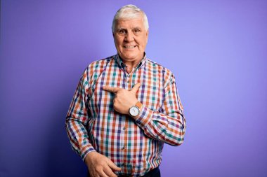 Kıdemli yakışıklı, beyaz gömlekli, soyutlanmış, mor arka planda neşeli, gülümseyen, mutlu ve doğal bir ifadeyle elini ve parmağını kaldırmış bir adam.