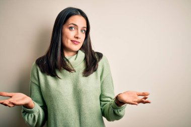 Mavi gözlü genç esmer kadın beyaz arka planda balıkçı yaka kazak giyiyor ve elleri ve kolları havada, kafası karışmış bir ifade takınıyor. Şüphe kavramı.