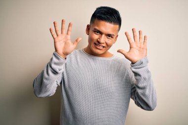 İzole edilmiş beyaz arka planda dikilen gündelik süveter giyen genç ve yakışıklı Latin adam on numarayı gösterirken kendinden emin ve mutlu bir şekilde gülümsüyor..