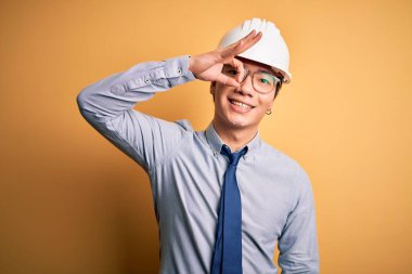 Güvenlik kaskı takan ve sarı arka planda kravat takan genç yakışıklı mimar el hareketleriyle gülümsüyor ve mutlu yüzle parmaklarının arasından bakıyor..