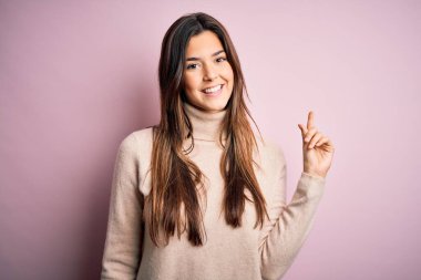 Balıkçı yaka kazak giyen genç ve güzel bir kız izole edilmiş pembe arka plan üzerinde duruyor ve bir numaralı parmağını gösterirken kendine güvenen ve mutlu bir şekilde gülümsüyor..