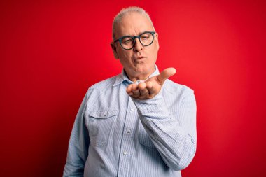Middle age handsome hoary man wearing casual striped shirt and glasses over red background looking at the camera blowing a kiss with hand on air being lovely and sexy. Love expression. clipart