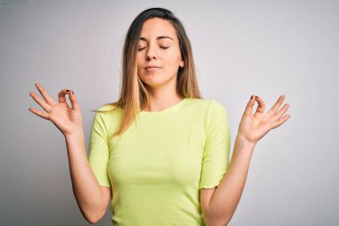 Mavi gözlü, yeşil tişörtlü, beyaz arka planda güzel sarışın kadın rahatla ve gözleri kapalı, parmaklarıyla meditasyon hareketi yaparken gülümse. Yoga konsepti.