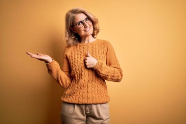 Orta yaşlı güzel sarışın kadın sarı arka planda günlük süveter ve gözlük takıyor avuç içini gösteriyor ve baş parmaklarıyla hareket ediyor, mutlu ve neşeli gülümsüyor.
