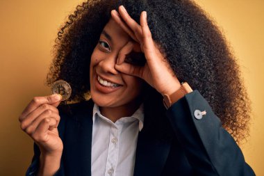 Afro saçlı, bitcoin 'i sarı arka planda tutan, gülümseyen, el pençe divan duran Afro-Amerikalı genç bir iş kadını.