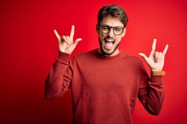 Sakallı, gözlüklü ve süveterli genç bir adam kırmızı arka planda dikilmiş deli gibi bağırıyor elleri havada rock sembolü yapıyor. Müzik yıldızı. Ağır müzik konsepti.