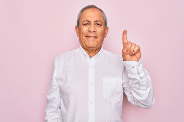 Üst düzey yakışıklı, gri saçlı, zarif tişörtlü, soyutlanmış pembe arka planda bir numarayı işaret eden, kendinden emin ve mutlu bir şekilde gülümseyen adam..