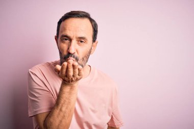 Orta yaşlı, beyaz tişörtlü bir adam izole edilmiş pembe arka planda dikilip kameraya bakarak havada el ele öpüşerek güzel ve seksi görünmeye çalışıyor. Aşk ifadesi.
