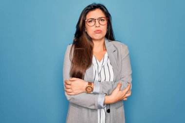 Genç İspanyol iş kadını mavi izole edilmiş arka planda duran gözlük takıyor. Yüzünde üzgün ve şok ifadesiyle kış soğuğundan titriyor ve donuyor.